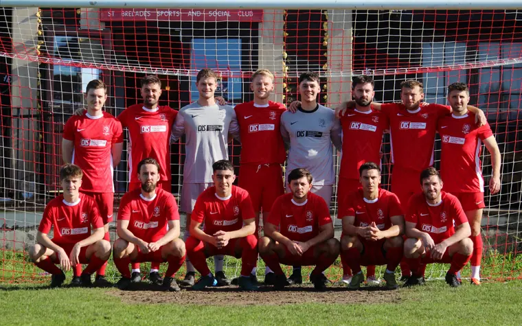 The St Brelade FC team in red in front of the goal, the clubhouse behind